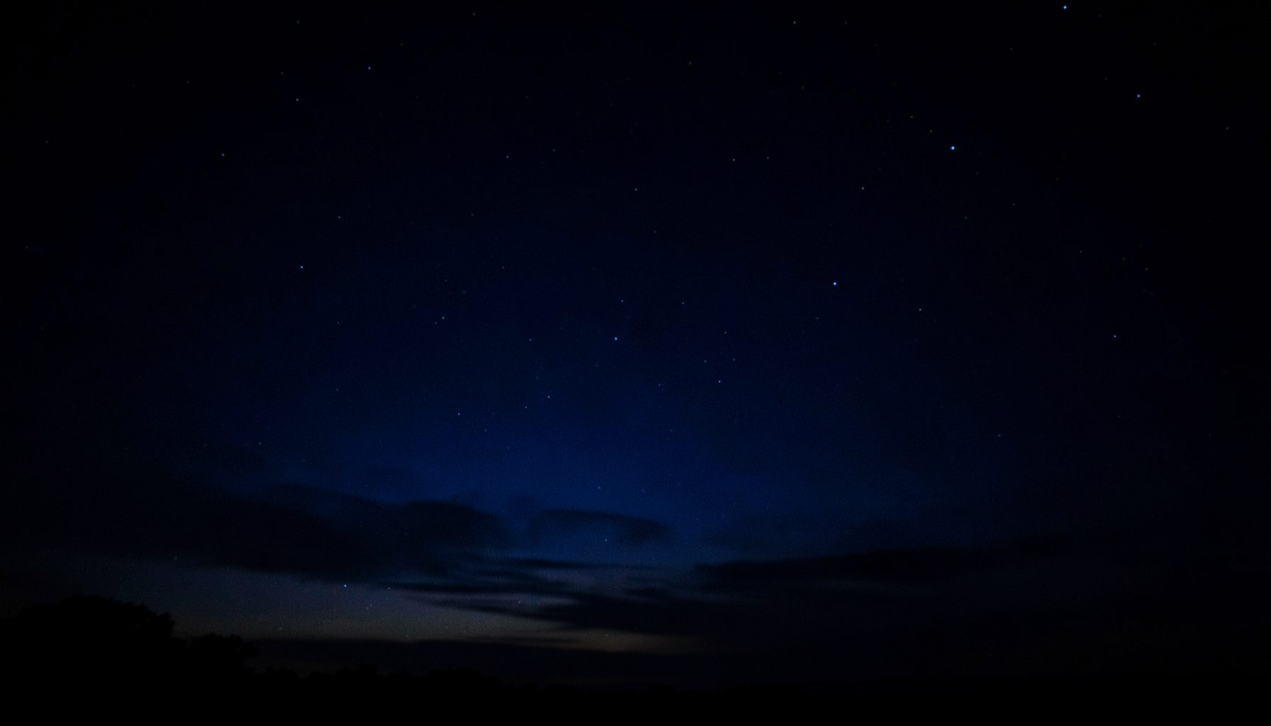 a dark low-key photograph of a deep indigo night sky scattered with faint stars and a soft glow near the horizon, moody and quiet